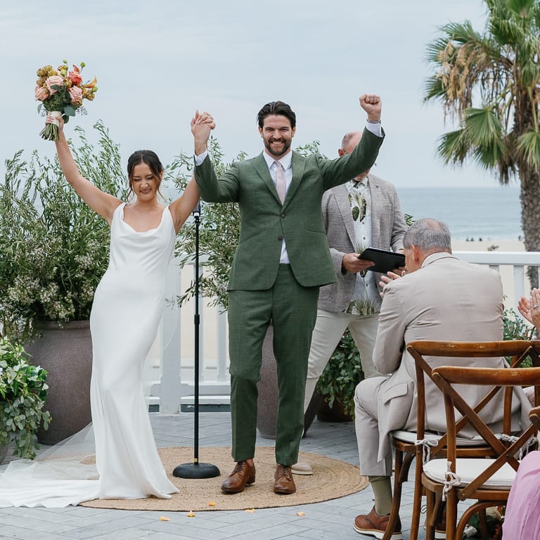 Bride in sleek slip gown and groom in green suit at altar on patio overlooking Santa Monica beach