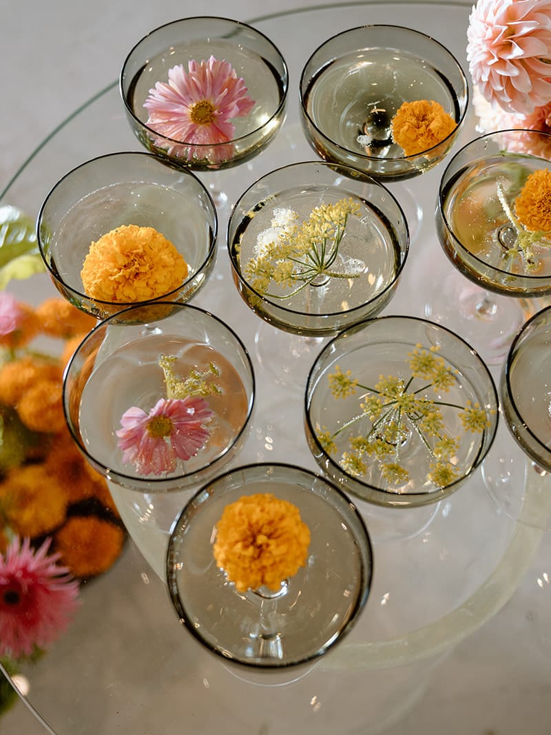 Champagne coupes decorated with floating pink and orange flowers on a glass table surrounded by colorful wedding florals.