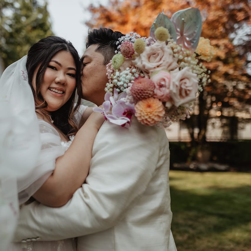 Bride and groom embrace with veil blowing in wind on the grounds of Rock Creek Gardens
