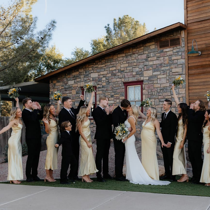 Bride and Groom share a kiss at rustic venue cheered on by their bridesmaids and groomsmen