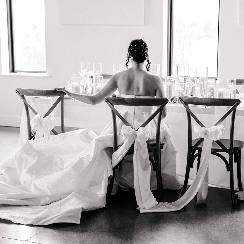 Black and white photo of a model wearing a bridal gown sitting at a table with bow detail